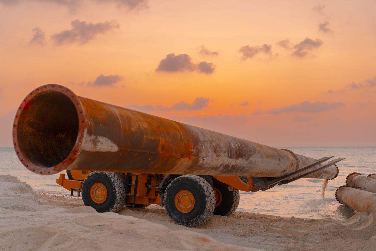 A rusted pipeline and machinery at a coastal construction site during sunset.